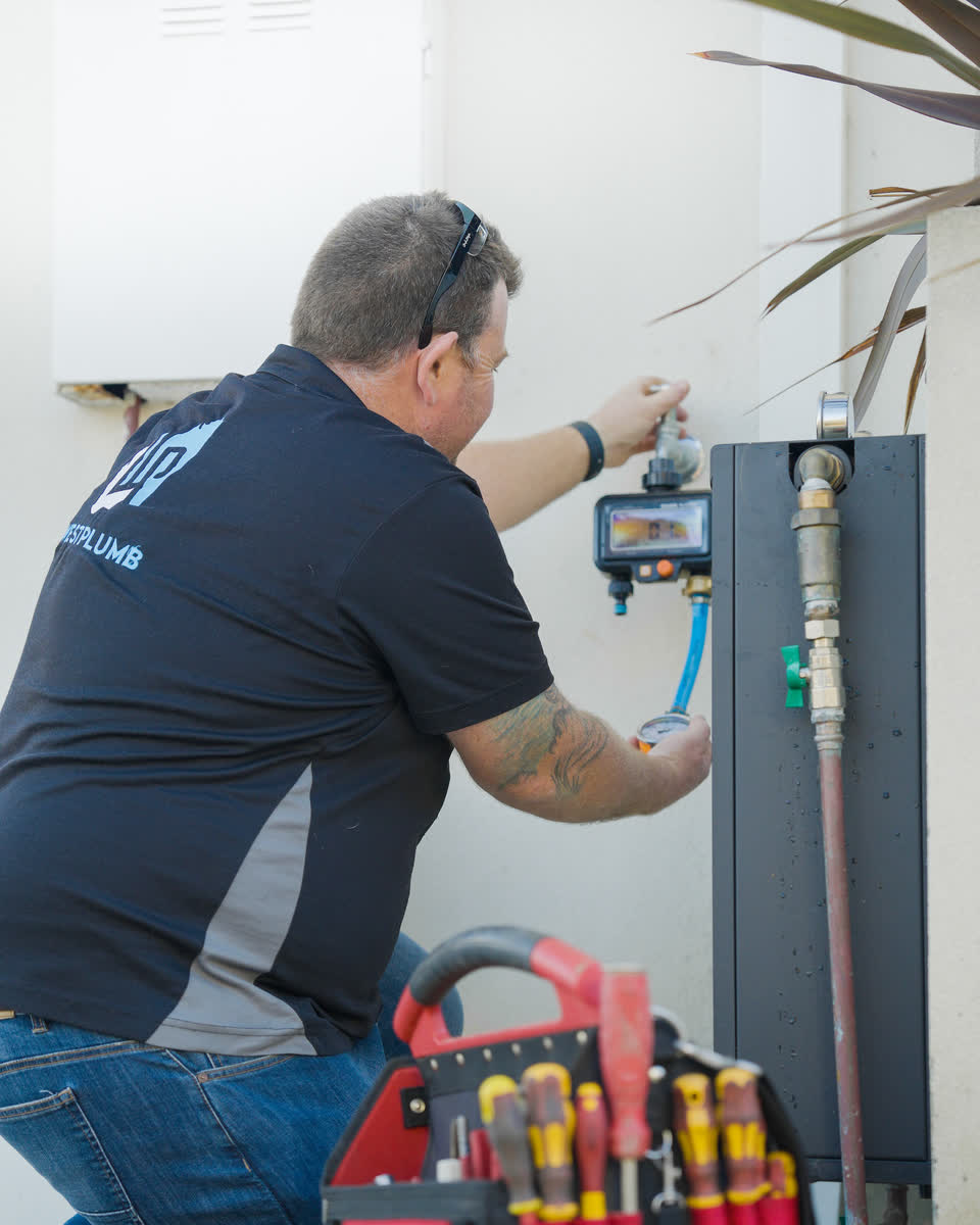 A Westplumb plumber kneeling at a hot water unit with his tool bag